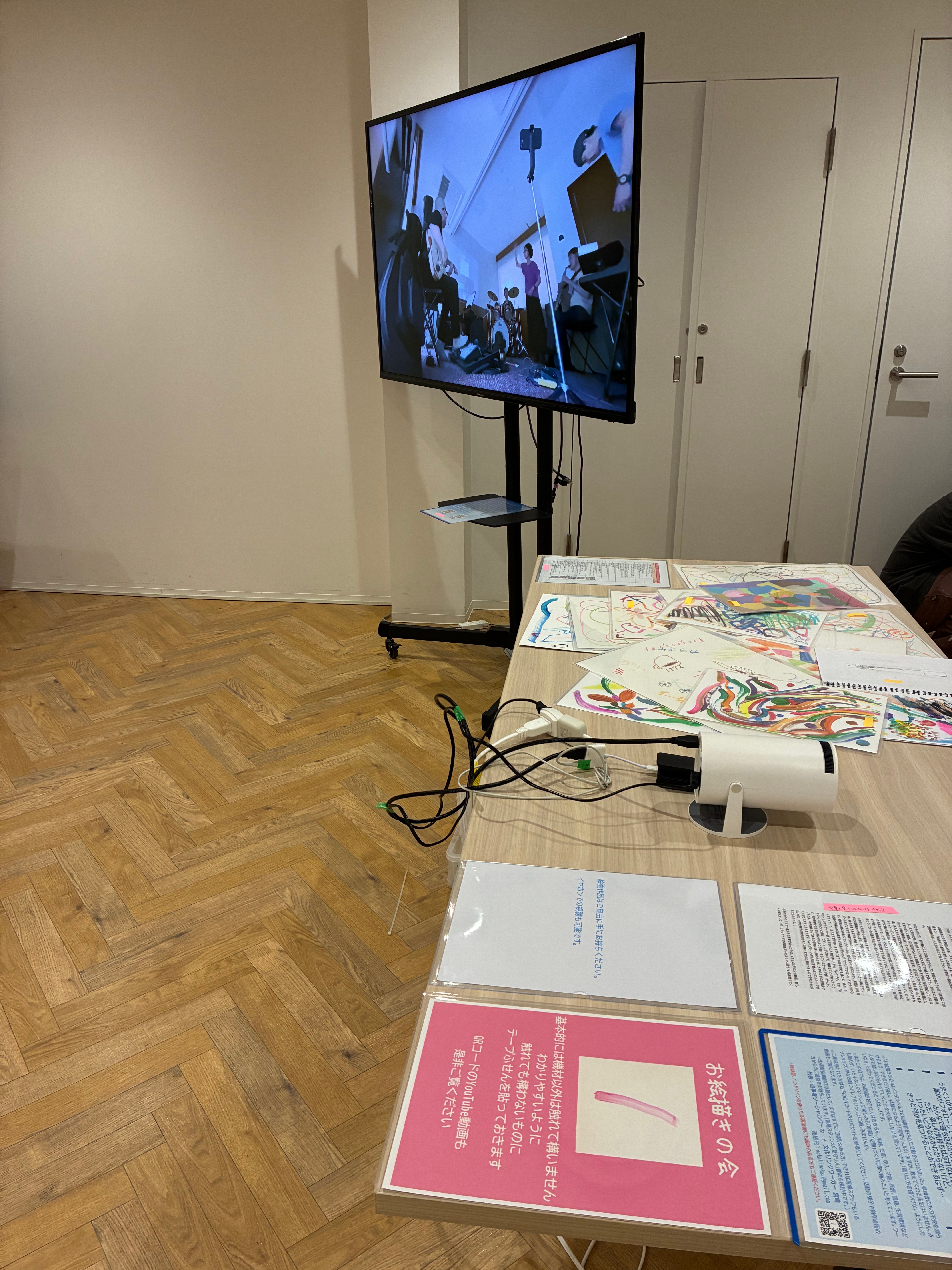 持ち込んだ作品の設置は一通り済ませた後に撮影した写真。アドバイスを受け、説明文などは、手前にあるテーブルの、右側に両面テープで留め、真ん中にプロジェクター、左に紙作品を置いた。テーブルの奥に大画面モニターがあり、試しの映像が映っている。