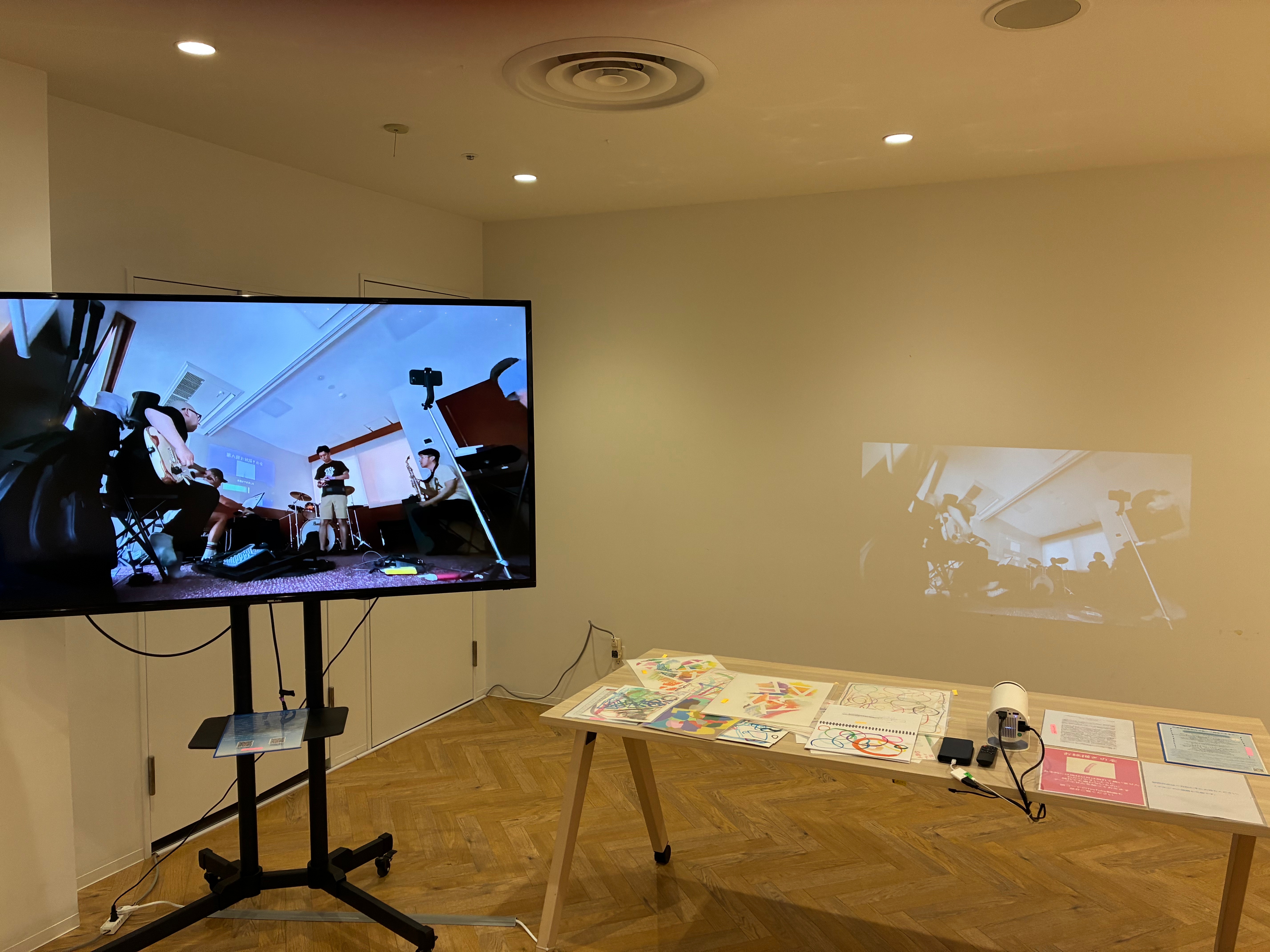 JamJam展の展示の全体像を写真に写した。左側に大画面で小ボリュームでお絵描きの会の作品の創作のプロセスを写しながら劇伴のように音づけしたものを編集した映像作品が写っている。右のテーブルの上には、左から紙作品、iPhoneやプロジェクター、右に説明文がある。プロジェクターには、大画面に映っている動画の編集をあまりしないで1時間ほどの作品となっているものが映っている。こちらはイヤホンで聴くようになっている。